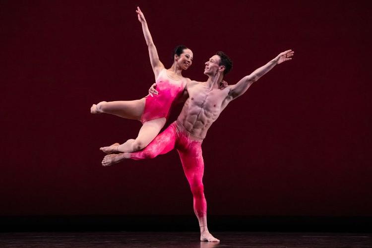 A dancer in hot pink pants lifting another dancer in a matching leotard on his side.